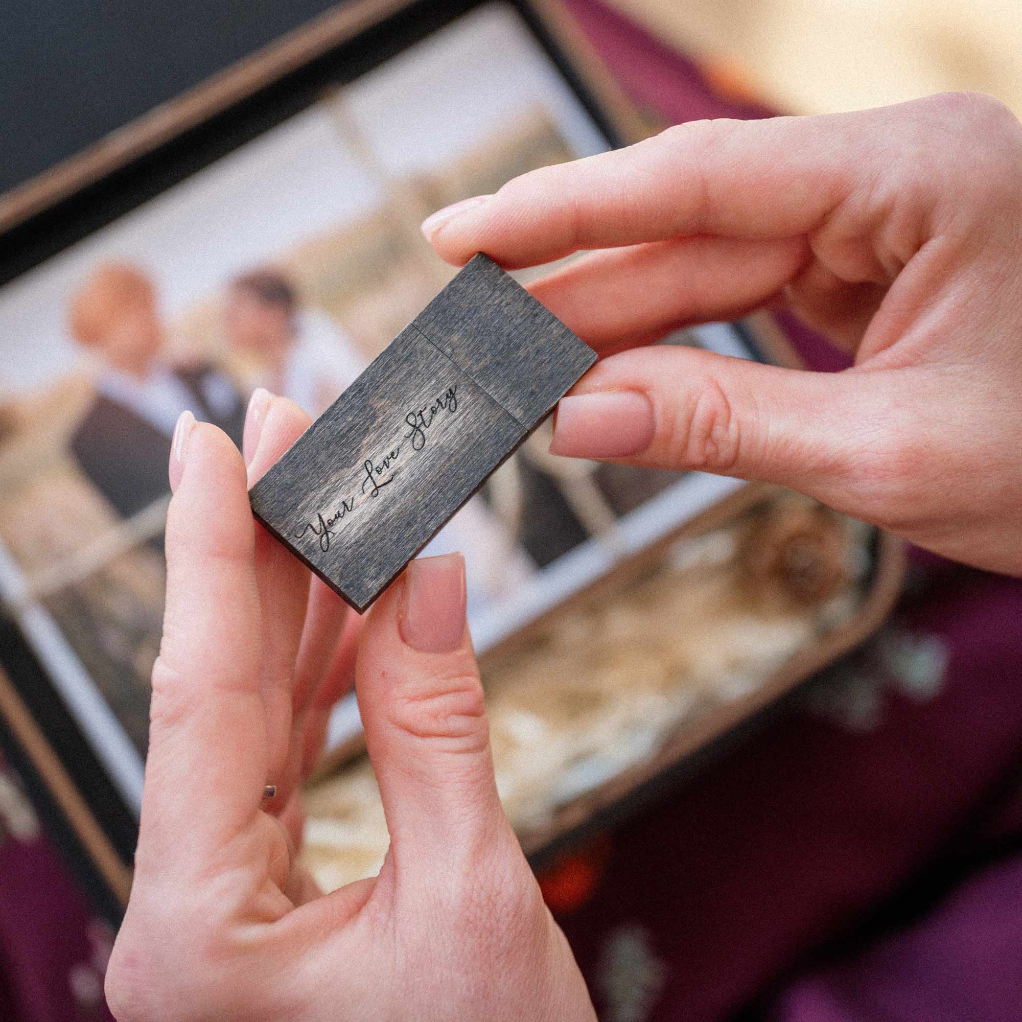 Hands holding engraved wooden USB drive above open black wedding photo box