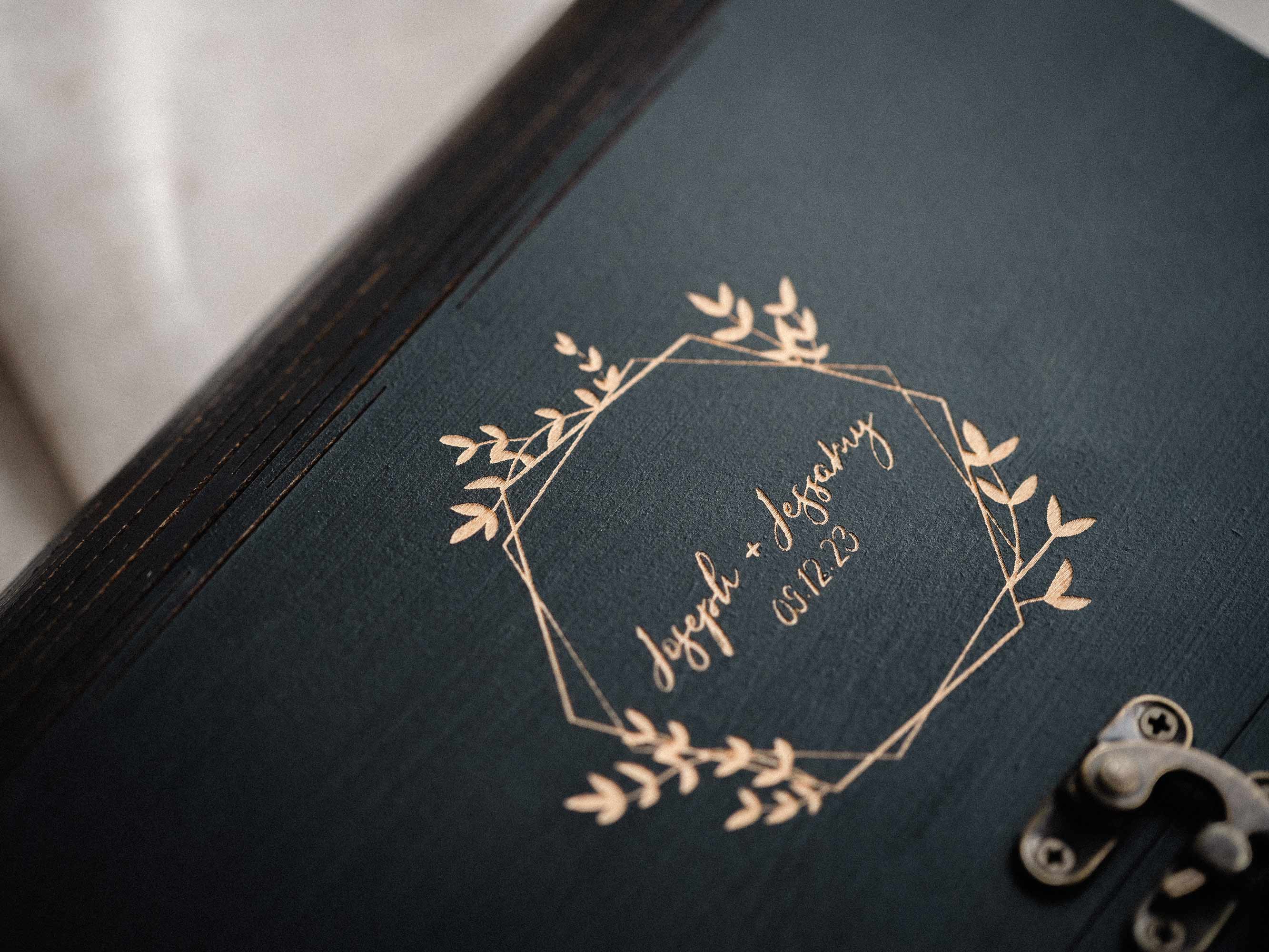 Close-up of engraved wreath personalization on black wooden photo box lid, wedding keepsake detail