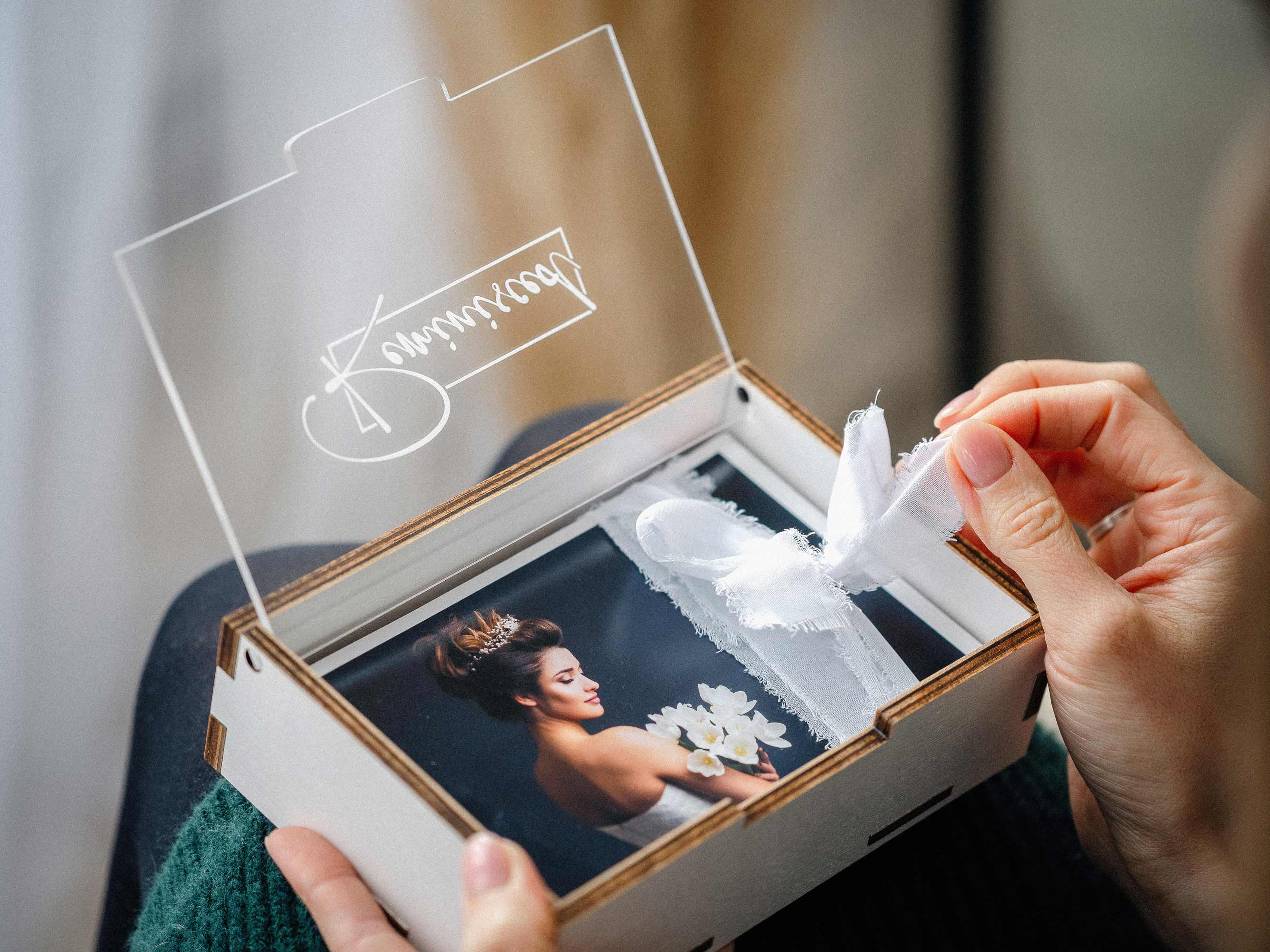 White wooden photo box with engraved acrylic lid and ribbon, 4x6 prints in hands.
