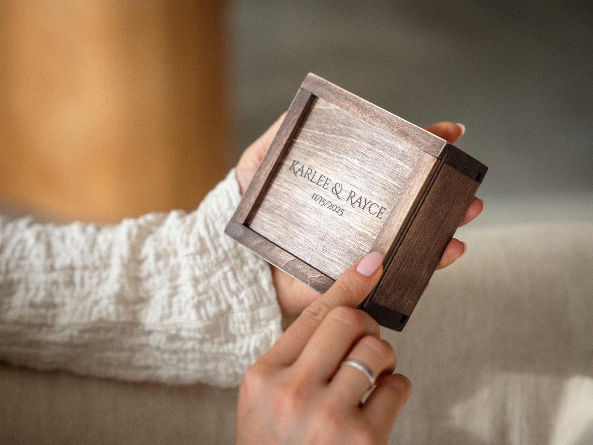Personalized walnut-style wooden USB box with engraved couple names, photographed in hands against a soft neutral background.